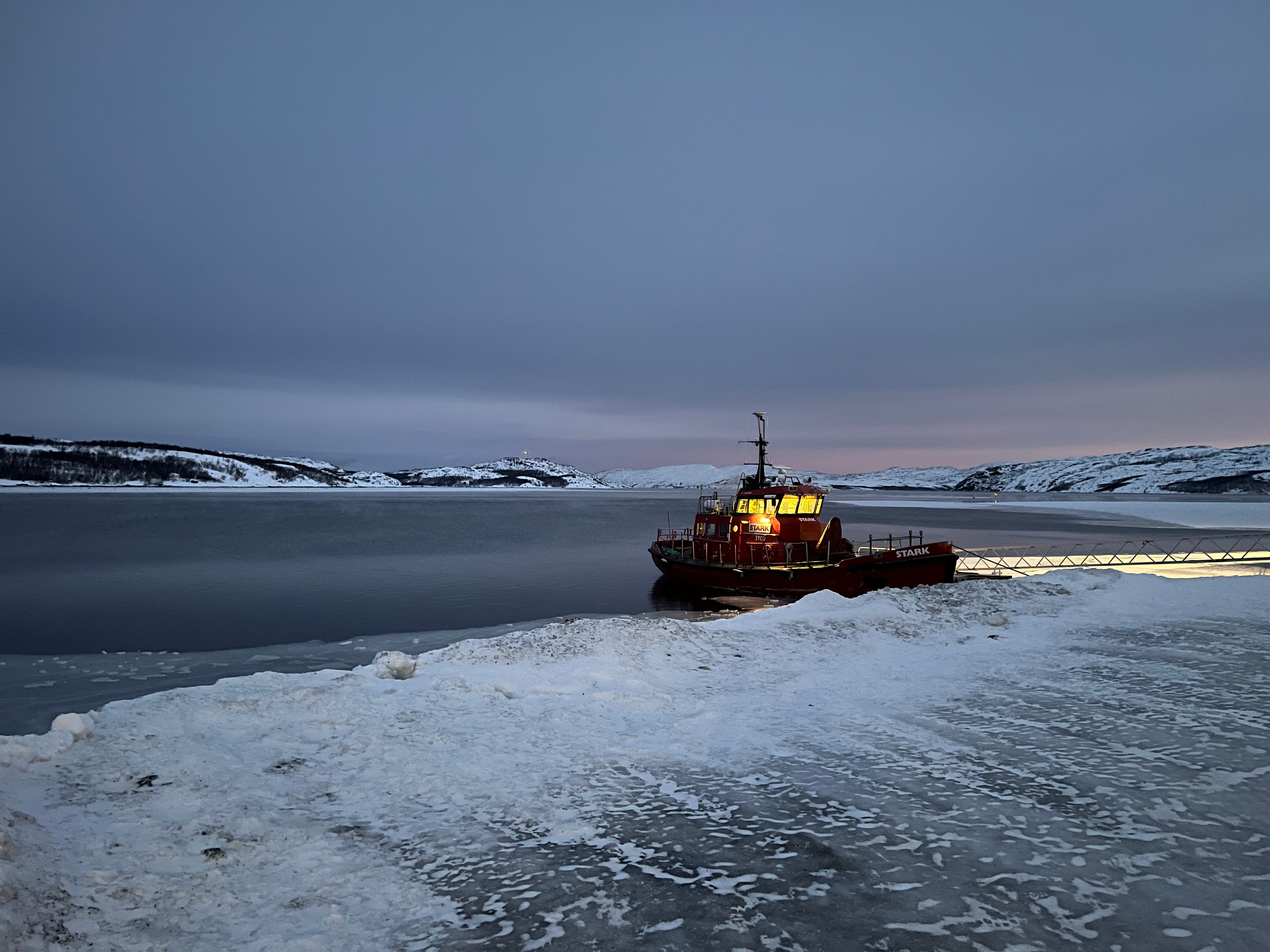 A boat sits docked on a icy, snow-covered shore, snowy hills in the distance, and the sky overcast and still dark, with only a hint of dawn in the distance