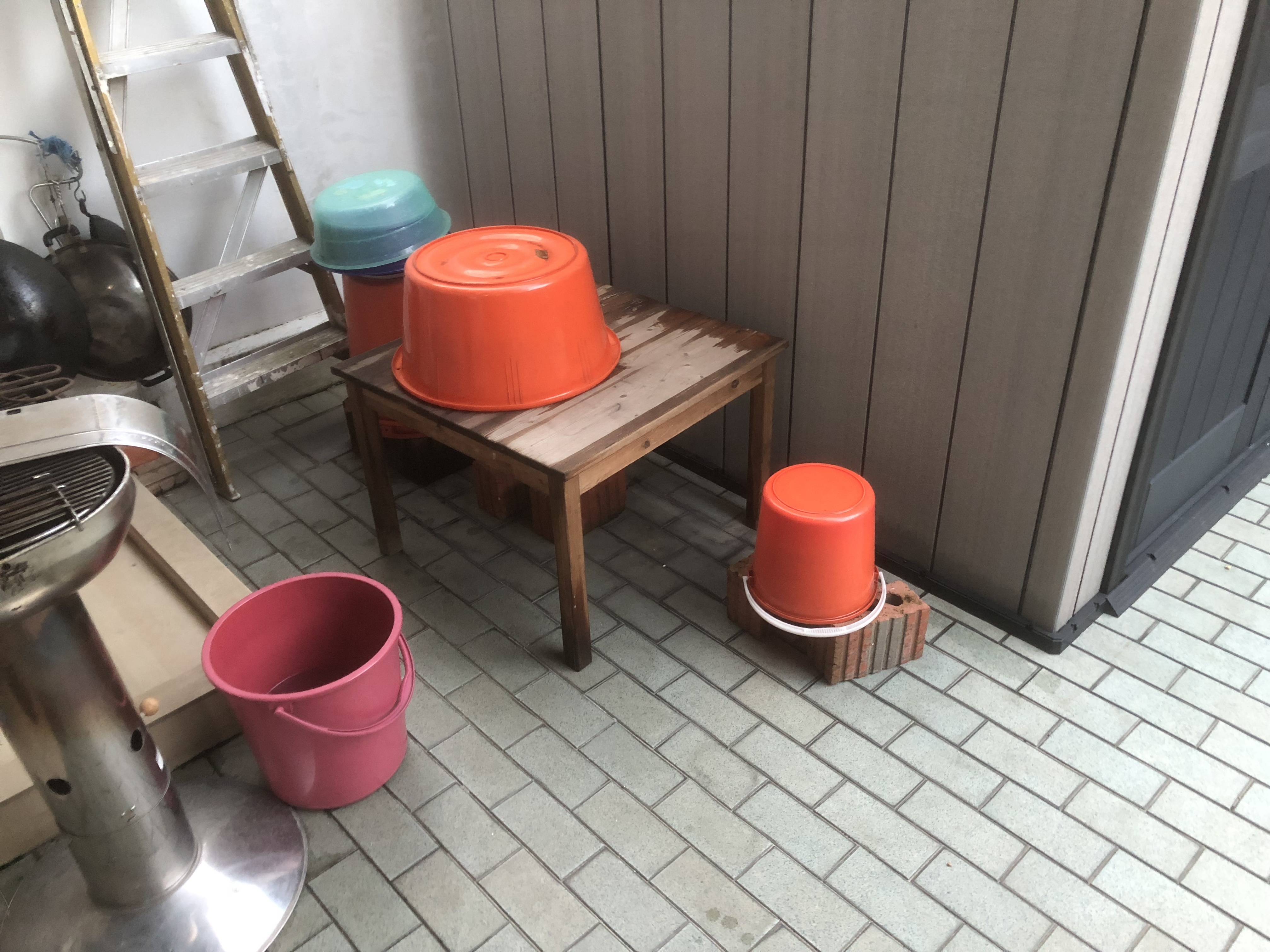 A square wooden table stands outdoors, next to a small shed, on blue ceramic tiles. Rest on and near the table are several pails