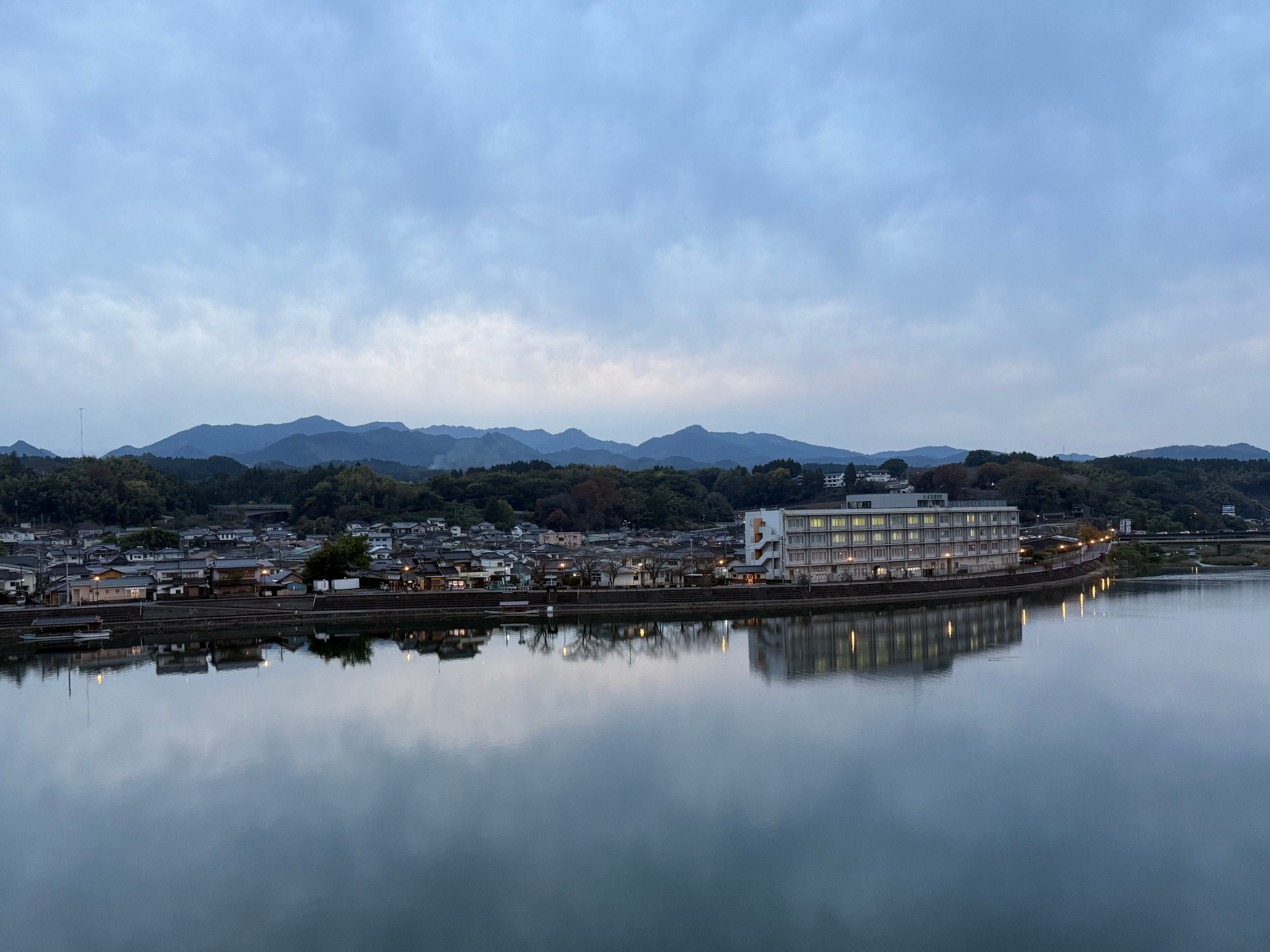 A rectangular, low-rise building sits amidst a neighbourhood of short, 1- to 2-storey buildings with traditional Japanese tiled roofs. In the foreground, a river, its water completely still, reflecting the evening sky above. In the distance, mountains